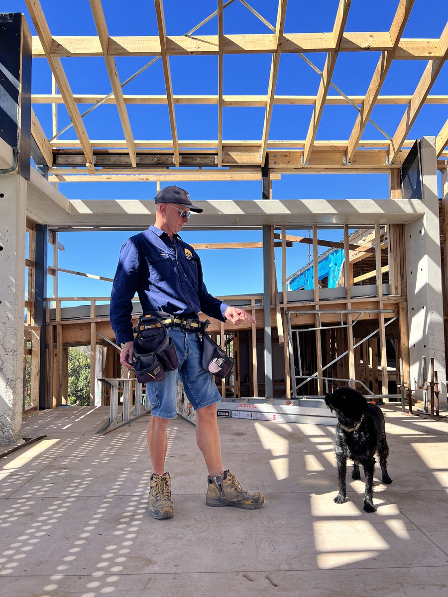 Steve on a residential build, standing under an exposed timber roof frame with his tool belt on and his dog beside him.